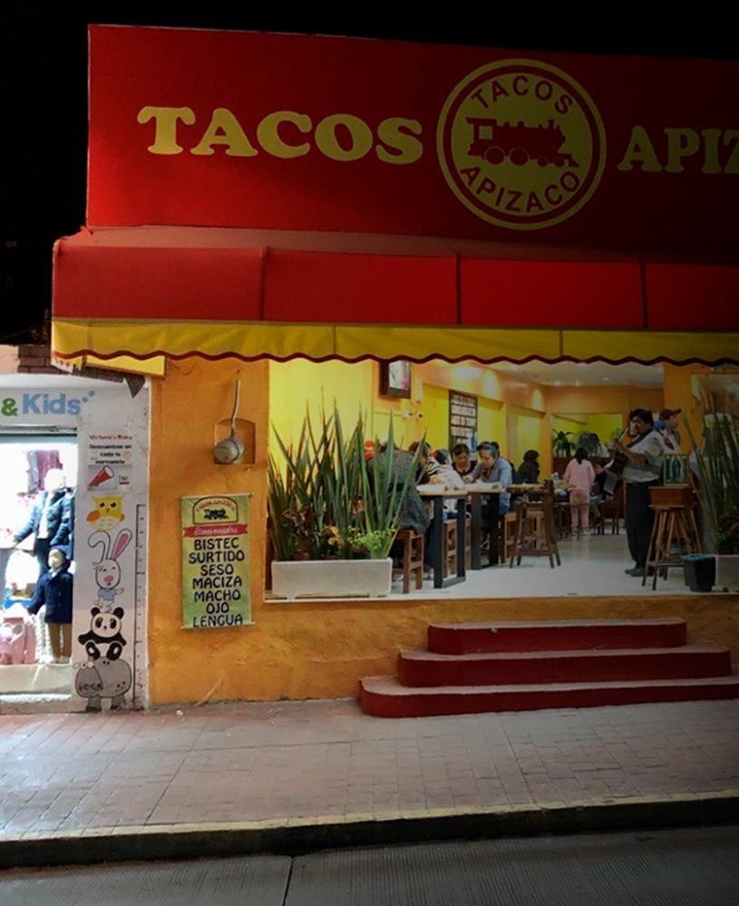 La misma fachada de noche, con el local iluminado y lleno de gente; a la entrada, el cartel con la lista: bistec, surtido, seso, maciza, macho, ojo y lengua.