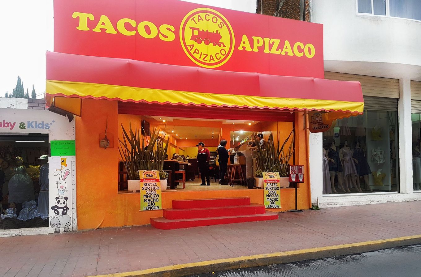 Fachada de Tacos Apizaco con toldo rojo y amarillo y escalones rojos; a la entrada, dos carteles con la lista: bistec, surtido, seso, maciza, macho, ojo y lengua.